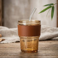 Clear glass tumbler with brown leather strap and glass straw on a wooden surface.