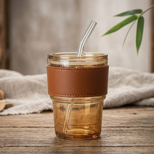 Clear glass tumbler with brown leather strap and glass straw on a wooden surface.